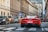A vibrant red sports car with a visible license plate is driving down a city street. Several other vehicles, including black and brown cars, are seen alongside. The street is lined with tall historic buildings. A few pedestrians stand near the sidewalk, with one holding a bag. Various traffic signs are visible, pointing and indicating directions.