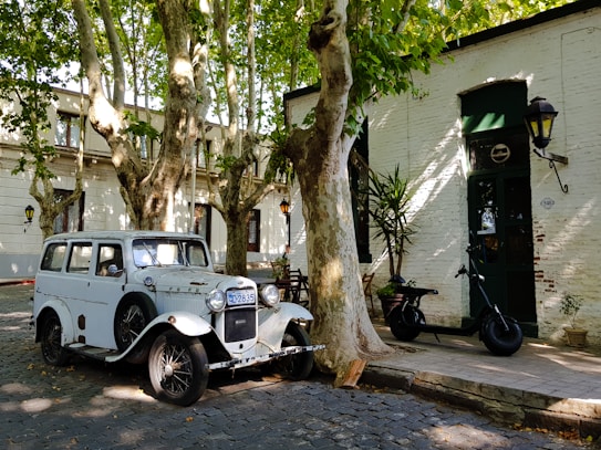 A vintage white car is parked on a cobblestone street under tall, leafy trees. Nearby is a classic white brick building with dark green accents, featuring large windows and a door. An old-style street lamp is attached to the wall, and a black scooter is parked on the sidewalk.