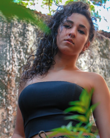 A young woman with curly hair and a nose ring, wearing a strapless black top, stands outdoors surrounded by greenery. She gazes directly at the camera with a neutral expression. The background features blurred foliage and a textured wall.