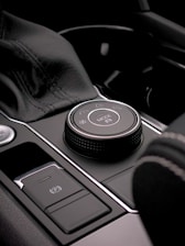 A close-up view of a car's interior control panel featuring a mode selection knob with icons indicating various settings. The area is composed of smooth, sleek materials with a metallic finish, including stitched leather and shiny chrome detailing.