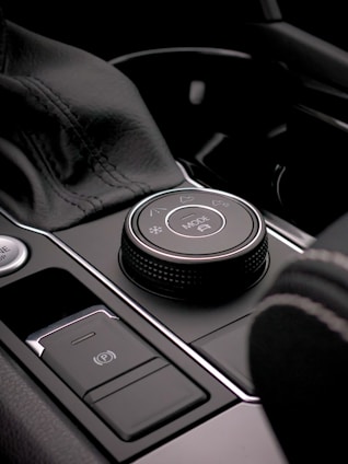 A close-up view of a car's interior control panel featuring a mode selection knob with icons indicating various settings. The area is composed of smooth, sleek materials with a metallic finish, including stitched leather and shiny chrome detailing.