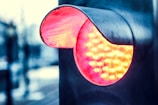 A close-up of a modern LED traffic signal, illuminated with a bright red light. The metallic casing reflects ambient light, giving a glossy appearance. The background appears blurred, suggesting an urban setting.