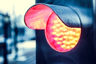 A close-up of a modern LED traffic signal, illuminated with a bright red light. The metallic casing reflects ambient light, giving a glossy appearance. The background appears blurred, suggesting an urban setting.