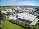 An aerial view of an industrial area featuring large warehouse buildings surrounded by greenery and residential areas. The scene includes parking lots with cars, roads, and nearby fields stretching out to the horizon. The weather appears clear and sunny, enhancing the visibility of the landscape and structures.