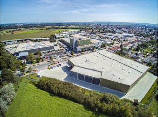 An aerial view of an industrial area featuring large warehouse buildings surrounded by greenery and residential areas. The scene includes parking lots with cars, roads, and nearby fields stretching out to the horizon. The weather appears clear and sunny, enhancing the visibility of the landscape and structures.