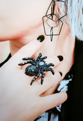 A close-up of a person's hand adorned with a large black spider ring. The fingernails are painted black, matching the ring and complementing the overall aesthetic. The image includes part of the face with a geometric earring, and strands of white or grey hair can be seen. The image conveys a gothic or edgy style.