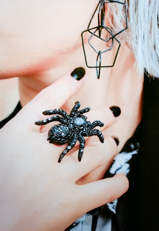 A close-up of a person's hand adorned with a large black spider ring. The fingernails are painted black, matching the ring and complementing the overall aesthetic. The image includes part of the face with a geometric earring, and strands of white or grey hair can be seen. The image conveys a gothic or edgy style.