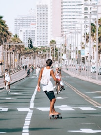 A young person skateboards down a sunny urban street lined with palm trees and tall buildings. Several other people are around, some riding bicycles, creating a relaxed, active atmosphere. The perspective is from behind the skateboarder, focusing on movement and leisure activities in a city setting.