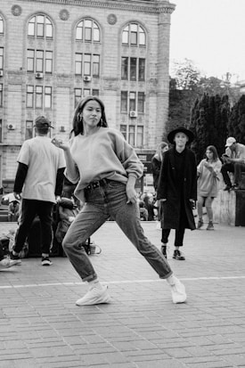 A person wearing a sweater and jeans performs a dance move in an urban outdoor setting. Behind, several people are casually gathered near a wall, and an old building with tall windows serves as the backdrop.