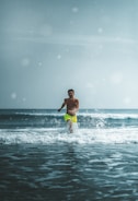 A person wearing bright yellow shorts runs through shallow ocean water, with waves crashing around them under a clear sky.