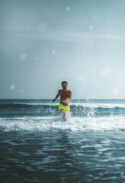 A person wearing bright yellow shorts runs through shallow ocean water, with waves crashing around them under a clear sky.