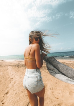 A woman with long hair and sunglasses is standing on a sandy beach, wearing a brown bikini top and light blue denim shorts. She is holding a patterned blanket or towel, with the ocean visible in the background under a partly cloudy sky.