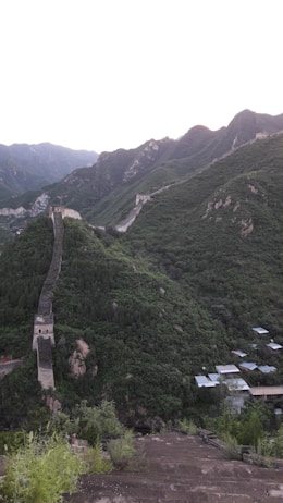 A section of the Great Wall of China winds across lush green mountains, showcasing ancient stone towers and fortifications. The landscape is vast, with a series of hills extending into the distance. Small structures are nestled at the base of the mountain.