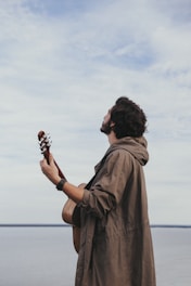 A person with curly hair holding an acoustic guitar appears to be gazing thoughtfully at the sky. They are wearing a long, brown coat and stand against a backdrop of a calm, expansive body of water and a cloudy sky.