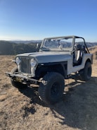 A rugged, off-road vehicle with a vintage design is parked on dry terrain. The Jeep, with large tires and an open top, is positioned in a hilly landscape under a clear blue sky. The background features rolling hills and sparse vegetation.