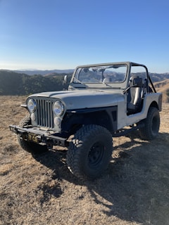 A rugged, off-road vehicle with a vintage design is parked on dry terrain. The Jeep, with large tires and an open top, is positioned in a hilly landscape under a clear blue sky. The background features rolling hills and sparse vegetation.