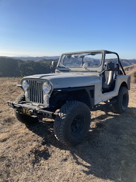 A rugged, off-road vehicle with a vintage design is parked on dry terrain. The Jeep, with large tires and an open top, is positioned in a hilly landscape under a clear blue sky. The background features rolling hills and sparse vegetation.