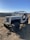 A rugged, off-road vehicle with a vintage design is parked on dry terrain. The Jeep, with large tires and an open top, is positioned in a hilly landscape under a clear blue sky. The background features rolling hills and sparse vegetation.