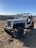 A rugged, off-road vehicle with a vintage design is parked on dry terrain. The Jeep, with large tires and an open top, is positioned in a hilly landscape under a clear blue sky. The background features rolling hills and sparse vegetation.