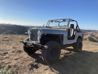 A rugged off-road vehicle, similar to a classic Jeep, is parked on a dry, grassy hilltop. The setting is mountainous with rolling hills and a clear blue sky in the background, suggesting a serene and adventurous outdoor atmosphere.