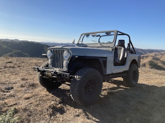 A rugged off-road vehicle, similar to a classic Jeep, is parked on a dry, grassy hilltop. The setting is mountainous with rolling hills and a clear blue sky in the background, suggesting a serene and adventurous outdoor atmosphere.