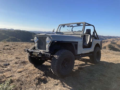 A rugged off-road vehicle, similar to a classic Jeep, is parked on a dry, grassy hilltop. The setting is mountainous with rolling hills and a clear blue sky in the background, suggesting a serene and adventurous outdoor atmosphere.