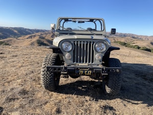 A rugged, vintage off-road vehicle with a prominent front grille and round headlights is parked on a dry, grassy hilltop. The landscape features rolling hills under a clear blue sky.