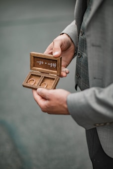 A person in a suit holds an open wooden box with two rings inside. The box lid has 'Mr. & Mrs.' written on it in cursive. The background is blurred, emphasizing the focus on the box and rings.