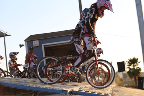 A group of young BMX riders wearing protective gear and helmets preparing to start a race on a BMX track. The front rider is poised on a ramp, ready to go. The scene is outdoors, captured during golden hour with a backdrop of a small building and a clear blue sky.