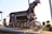 A group of young BMX riders wearing protective gear and helmets preparing to start a race on a BMX track. The front rider is poised on a ramp, ready to go. The scene is outdoors, captured during golden hour with a backdrop of a small building and a clear blue sky.