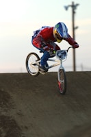 A person wearing protective gear, including a helmet and colorful clothing, is performing a BMX trick or race maneuver on a dirt track. The bike has a visible front number plate, and the rider is leaning forward intensely focused, suggesting high-speed action.