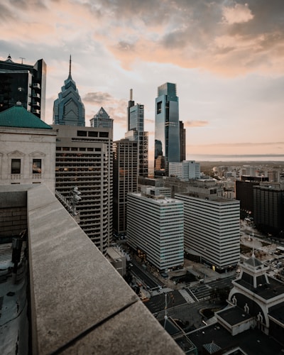 A cityscape featuring modern skyscrapers with varied architectural designs, captured during sunset. The clouds are tinged with warm colors of the setting sun, providing a soft glow over the high-rise buildings and streets below.