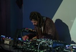 A person with long hair is intently focused on a table full of electronic music equipment, including mixers and synthesizers. The scene is dimly lit, suggesting a nightclub or studio setting, with shadows and colored lights casting an atmospheric backdrop.