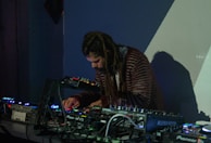 A person with long hair is intently focused on a table full of electronic music equipment, including mixers and synthesizers. The scene is dimly lit, suggesting a nightclub or studio setting, with shadows and colored lights casting an atmospheric backdrop.