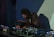 A person with long hair is intently focused on a table full of electronic music equipment, including mixers and synthesizers. The scene is dimly lit, suggesting a nightclub or studio setting, with shadows and colored lights casting an atmospheric backdrop.