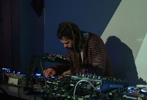 A person with long hair is intently focused on a table full of electronic music equipment, including mixers and synthesizers. The scene is dimly lit, suggesting a nightclub or studio setting, with shadows and colored lights casting an atmospheric backdrop.