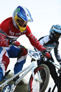 Two BMX bikers are in mid-action, wearing colorful gear and helmets. One rider, dressed in red, white, and blue, is prominently in the foreground, gripping the handlebars of a white bike with an intense focus. The background is slightly blurred, giving a sense of speed and motion.