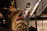 A close-up view of a brass musical instrument, possibly a tuba, with intricate reflections on its shiny surface. Several music stands with sheet music are positioned to the side, and there are blurred figures of musicians in the background.