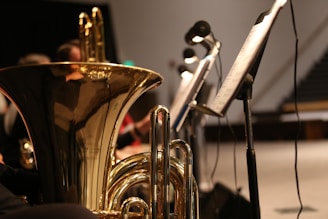 A close-up view of a brass musical instrument, possibly a tuba, with intricate reflections on its shiny surface. Several music stands with sheet music are positioned to the side, and there are blurred figures of musicians in the background.