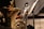 A close-up view of a brass musical instrument, possibly a tuba, with intricate reflections on its shiny surface. Several music stands with sheet music are positioned to the side, and there are blurred figures of musicians in the background.