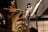 A close-up view of a brass musical instrument, possibly a tuba, with intricate reflections on its shiny surface. Several music stands with sheet music are positioned to the side, and there are blurred figures of musicians in the background.