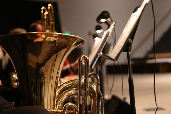 A close-up view of a brass musical instrument, possibly a tuba, with intricate reflections on its shiny surface. Several music stands with sheet music are positioned to the side, and there are blurred figures of musicians in the background.