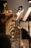 A close-up view of a brass musical instrument, possibly a tuba or euphonium, with sheet music on stands in the background. The setting appears to be a stage or rehearsal space, with musicians partially visible. The lighting is soft, focused on the instruments and the music stands.