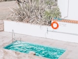 A small swimming pool with clear blue water is surrounded by a tiled patio. There is a pool ladder on one side and a vivid orange life ring hanging on the wall nearby, next to a sign indicating the shallow end as 3ft or 0.9m deep. Tall, spiky plants frame the area, adding a touch of greenery.