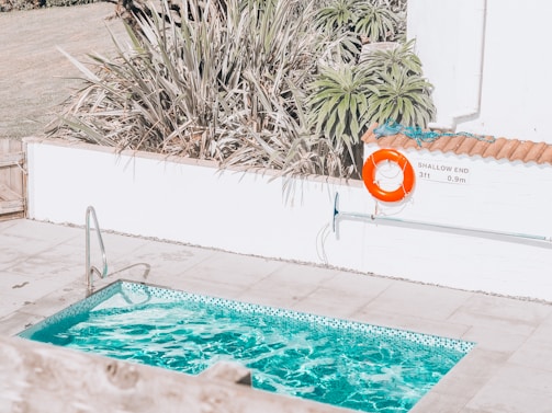 A small swimming pool with clear blue water is surrounded by a tiled patio. There is a pool ladder on one side and a vivid orange life ring hanging on the wall nearby, next to a sign indicating the shallow end as 3ft or 0.9m deep. Tall, spiky plants frame the area, adding a touch of greenery.