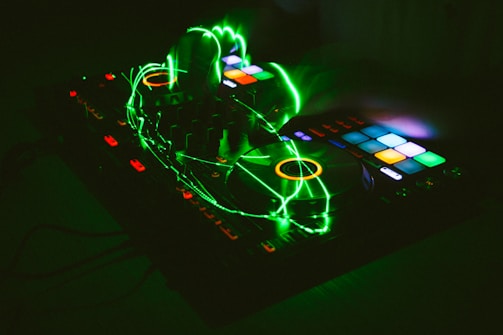 A DJ controller with colorful backlit buttons and turntables, accented by vibrant green light trails. The image captures a dynamic and energetic atmosphere with hands manipulating the controls, suggesting a live performance or mixing session.