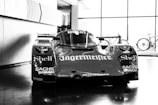 A vintage race car is prominently displayed in a showroom, featuring prominent sponsor logos such as Shell, Jägermeister, Sachs, and Bosch. The car exhibits a streamlined design with a glossy finish. Bicycles can be seen in the background near large windows that let in ample natural light, creating reflections on the polished floor.