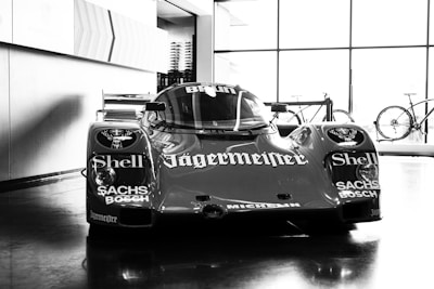 A vintage race car is prominently displayed in a showroom, featuring prominent sponsor logos such as Shell, Jägermeister, Sachs, and Bosch. The car exhibits a streamlined design with a glossy finish. Bicycles can be seen in the background near large windows that let in ample natural light, creating reflections on the polished floor.