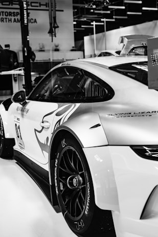 A sleek racing car is displayed in an indoor setting, featuring aerodynamic designs and decals. The car has a smooth, curvy body with clear sponsor logos and branding. The setting suggests a showroom or auto expo with other vehicles visible in the background.