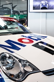 A close-up of a race car featuring prominent Mobil 1 branding on its hood. The car is white with blue and red text. It has a sleek headlight on the left side. In the background, there is a green object and another car with its hood open inside a garage or showroom. A large screen or display is visible, showing an image of a race car with the number 48 on it.